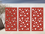 Metal privacy screen with a modern bubble pattern of varying circle sizes, finished in Signal Red powder coating, displayed outdoors against a white wall, providing both privacy and an artistic element to the space.