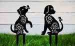 Decorative metal garden stakes featuring two sitting dogs with floral patterns, one with a butterfly on its nose, both finished in Night Sky Matt Black powder coating. The pieces are mounted in grass against a white wooden backdrop, adding a playful and artistic touch to any outdoor garden space.