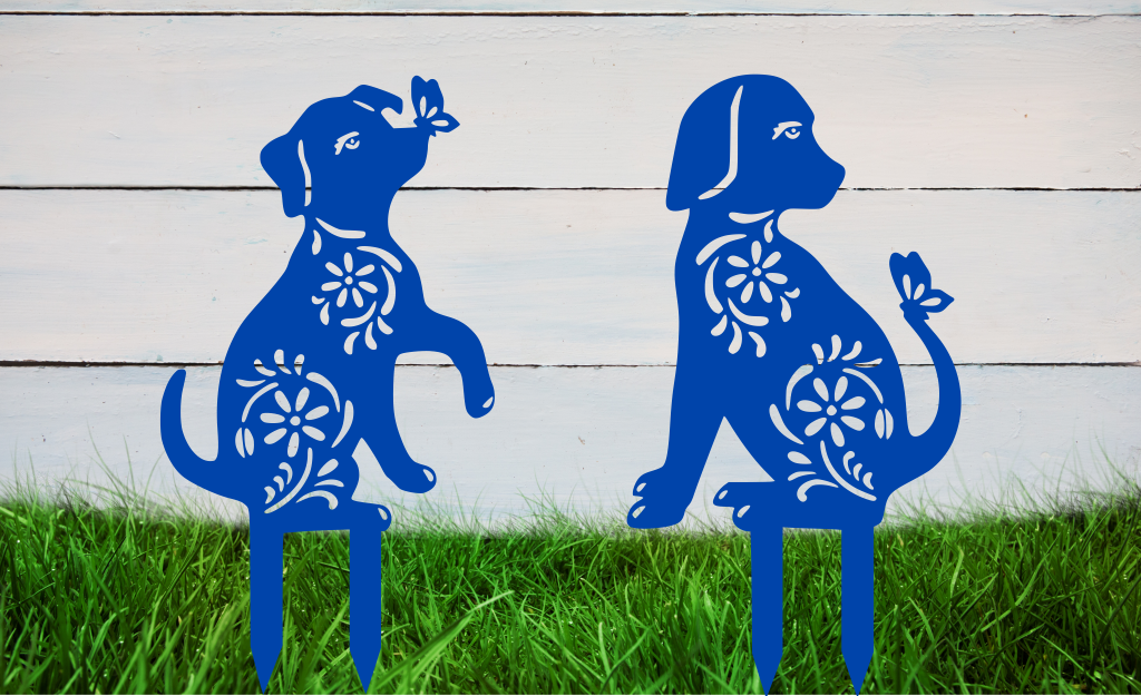 Decorative metal garden stakes featuring two sitting dogs with floral patterns, one with a butterfly on its nose, both finished in Blaze Blue Gloss powder coating. The pieces are mounted in grass against a white wooden backdrop, adding a playful and artistic touch to any outdoor garden space.