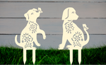 Decorative metal garden stakes featuring two sitting dogs with floral patterns, one with a butterfly on its nose, both finished in Canola Cream powder coating. The pieces are mounted in grass against a white wooden backdrop, adding a playful and artistic touch to any outdoor garden space.