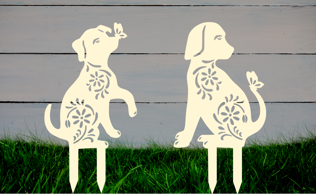 Decorative metal garden stakes featuring two sitting dogs with floral patterns, one with a butterfly on its nose, both finished in Canola Cream powder coating. The pieces are mounted in grass against a white wooden backdrop, adding a playful and artistic touch to any outdoor garden space.