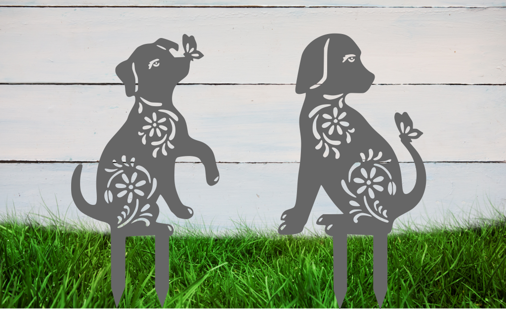 Decorative metal garden stakes featuring two sitting dogs with floral patterns, one with a butterfly on its nose, both finished in Berry Grey Gloss powder coating. The pieces are mounted in grass against a white wooden backdrop, adding a playful and artistic touch to any outdoor garden space.