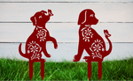 Decorative metal garden stakes featuring two sitting dogs with floral patterns, one with a butterfly on its nose, both finished in Manor Red powder coating. The pieces are mounted in grass against a white wooden backdrop, adding a playful and artistic touch to any outdoor garden space.