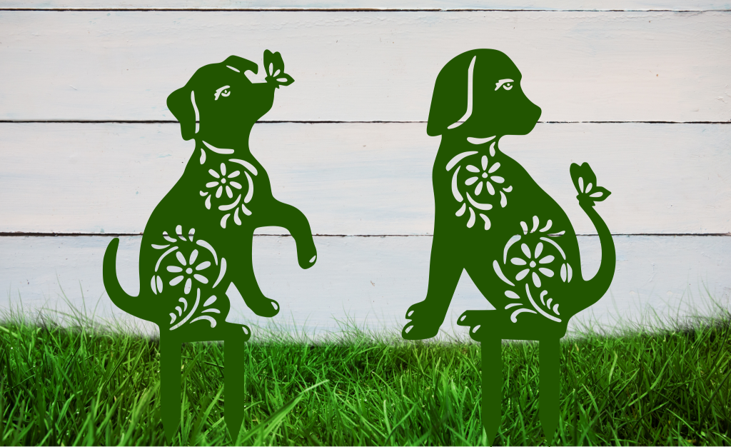 Decorative metal garden stakes featuring two sitting dogs with floral patterns, one with a butterfly on its nose, both finished in Mistletoe powder coating. The pieces are mounted in grass against a white wooden backdrop, adding a playful and artistic touch to any outdoor garden space.