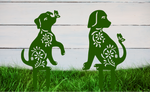 Decorative metal garden stakes featuring two sitting dogs with floral patterns, one with a butterfly on its nose, both finished in Mistletoe powder coating. The pieces are mounted in grass against a white wooden backdrop, adding a playful and artistic touch to any outdoor garden space.