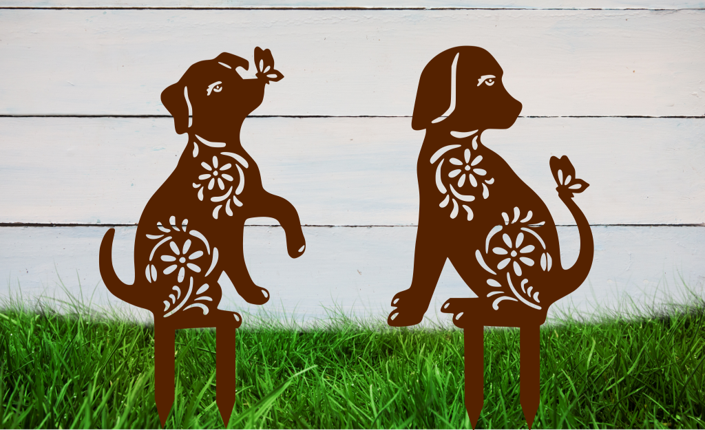 Decorative metal garden stakes featuring two sitting dogs with floral patterns, one with a butterfly on its nose, both finished in Rust Patina powder coating. The pieces are mounted in grass against a white wooden backdrop, adding a playful and artistic touch to any outdoor garden space