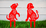 Decorative metal garden stakes featuring two sitting dogs with floral patterns, one with a butterfly on its nose, both finished in Signal Red powder coating. The pieces are mounted in grass against a white wooden backdrop, adding a playful and artistic touch to any outdoor garden space.