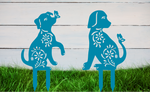 Decorative metal garden stakes featuring two sitting dogs with floral patterns, one with a butterfly on its nose, both finished in Wedgwood Satin powder coating. The pieces are mounted in grass against a white wooden backdrop, adding a playful and artistic touch to any outdoor garden space.