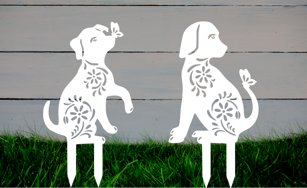 Decorative metal garden stakes featuring two sitting dogs with floral patterns, one with a butterfly on its nose, both finished in Bright White Gloss powder coating. The pieces are mounted in grass against a white wooden backdrop, adding a playful and artistic touch to any outdoor garden space.