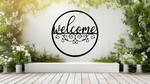 Metal wall art featuring a circular 'Welcome' sign with intricate scrollwork below the text, finished in Night Sky Matt Black powder coating, mounted on a white fence surrounded by lush greenery, adding a charming and inviting touch to an outdoor garden space.