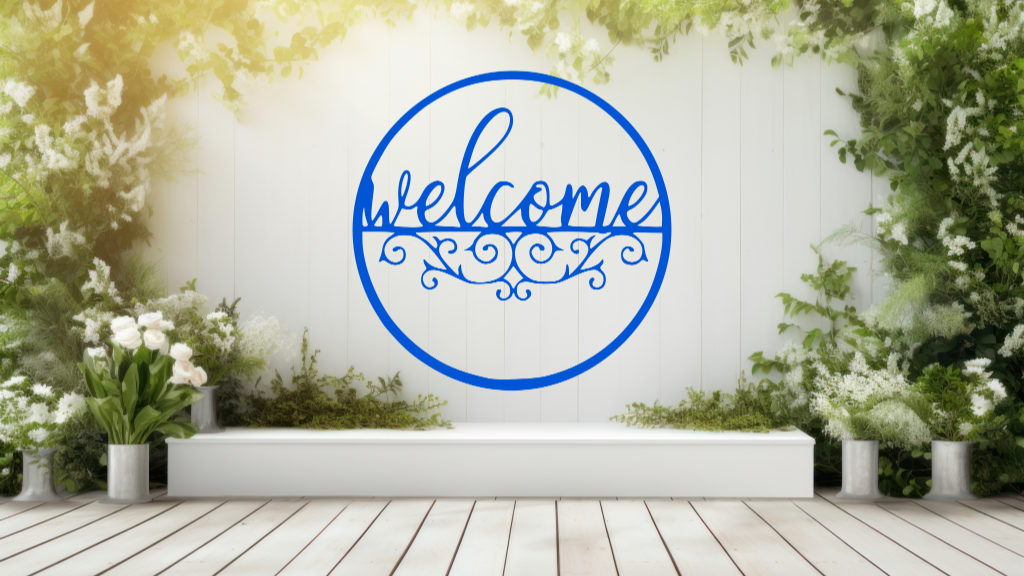 Metal wall art featuring a circular 'Welcome' sign with intricate scrollwork below the text, finished in Blaze Blue Gloss powder coating, mounted on a white fence surrounded by lush greenery, adding a charming and inviting touch to an outdoor garden space.