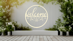 Metal wall art featuring a circular 'Welcome' sign with intricate scrollwork below the text, finished in Canola Cream powder coating, mounted on a white fence surrounded by lush greenery, adding a charming and inviting touch to an outdoor garden space.