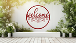 Metal wall art featuring a circular 'Welcome' sign with intricate scrollwork below the text, finished in Manor Red powder coating, mounted on a white fence surrounded by lush greenery, adding a charming and inviting touch to an outdoor garden space.