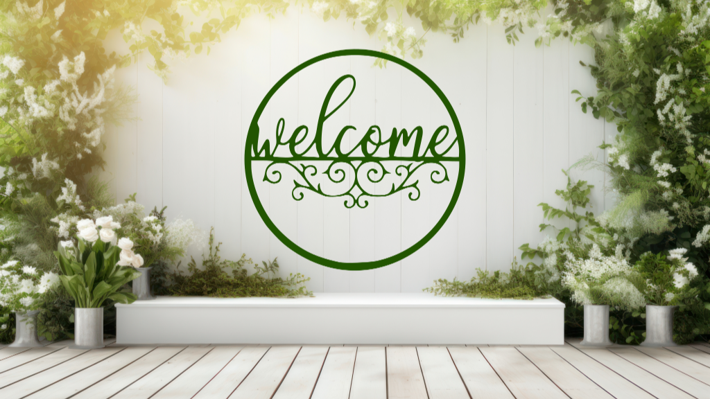 Metal wall art featuring a circular 'Welcome' sign with intricate scrollwork below the text, finished in Mistletoe powder coating, mounted on a white fence surrounded by lush greenery, adding a charming and inviting touch to an outdoor garden space.