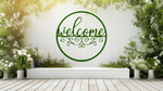 Metal wall art featuring a circular 'Welcome' sign with intricate scrollwork below the text, finished in Mistletoe powder coating, mounted on a white fence surrounded by lush greenery, adding a charming and inviting touch to an outdoor garden space.