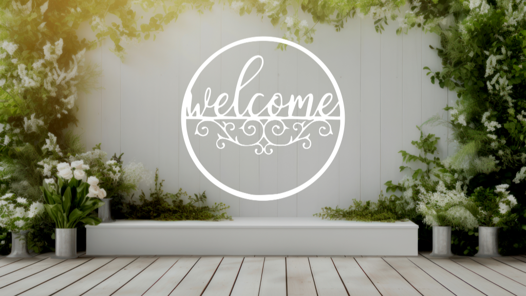 Metal wall art featuring a circular 'Welcome' sign with intricate scrollwork below the text, finished in Bright White Gloss powder coating, mounted on a white fence surrounded by lush greenery, adding a charming and inviting touch to an outdoor garden space.