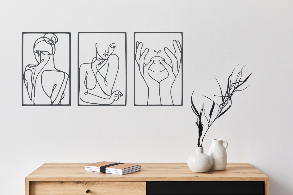 Metal wall art set featuring three minimalist line-drawing silhouettes of feminine figures in graceful poses, finished in Berry Grey Gloss powder coating, mounted on a white wall above a natural wood console, adding an elegant and artistic touch to modern interiors.