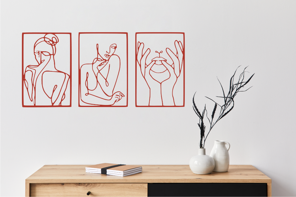 Metal wall art set featuring three minimalist line-drawing silhouettes of feminine figures in graceful poses, finished in Signal Red powder coating, mounted on a white wall above a natural wood console, adding an elegant and artistic touch to modern interiors.