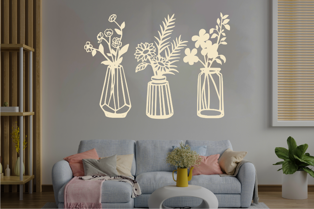 Set of three metal wall art pieces, each featuring a unique vase with blooming flowers, finished in Canola Cream powder coating. The artwork is displayed on a white wall above a cozy sofa, adding a delicate and artistic floral touch to the living room.