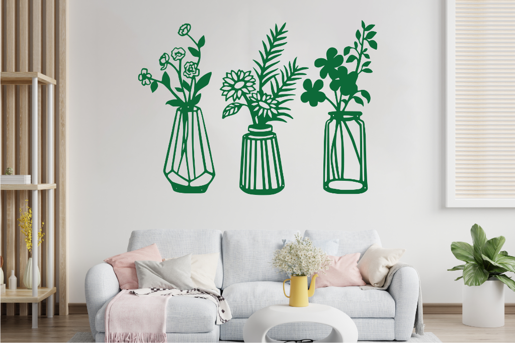 Set of three metal wall art pieces, each featuring a unique vase with blooming flowers, finished in Mistletoe powder coating. The artwork is displayed on a white wall above a cozy sofa, adding a delicate and artistic floral touch to the living room.