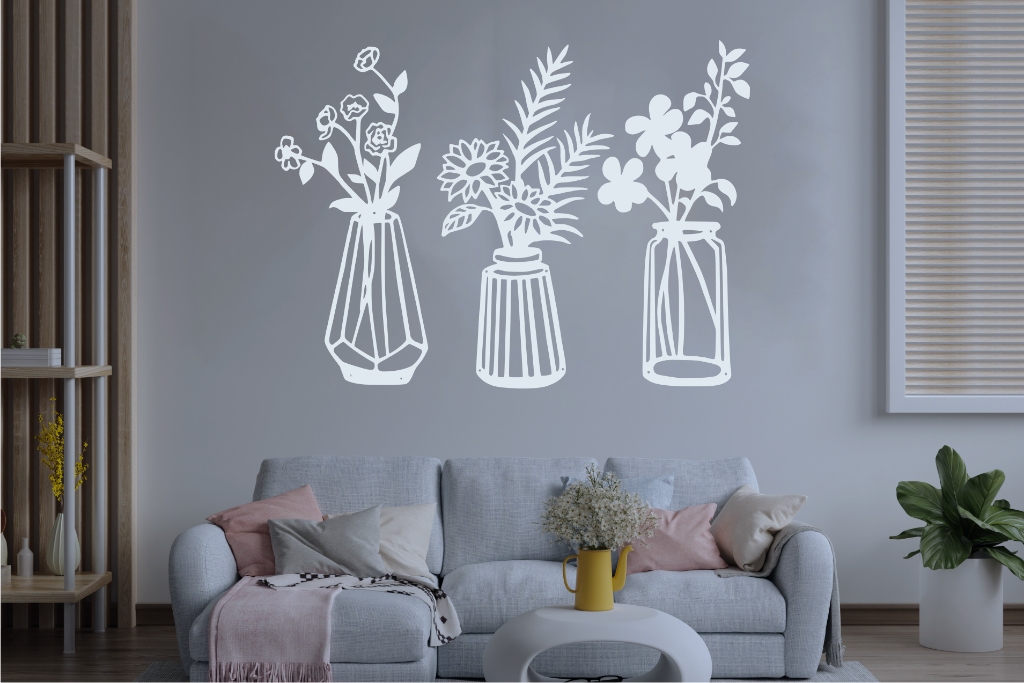 Set of three metal wall art pieces, each featuring a unique vase with blooming flowers, finished in Manor Red powder coating. The artwork is displayed on a white wall above a cozy sofa, adding a delicate and artistic floral touch to the living room.