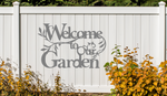 Metal wall art sign featuring the phrase 'Welcome to our Garden' with decorative branches and leaves integrated into the design, finished in Berry Grey Gloss powder coating. The sign is mounted on a white fence, adding a welcoming touch to an outdoor garden space.