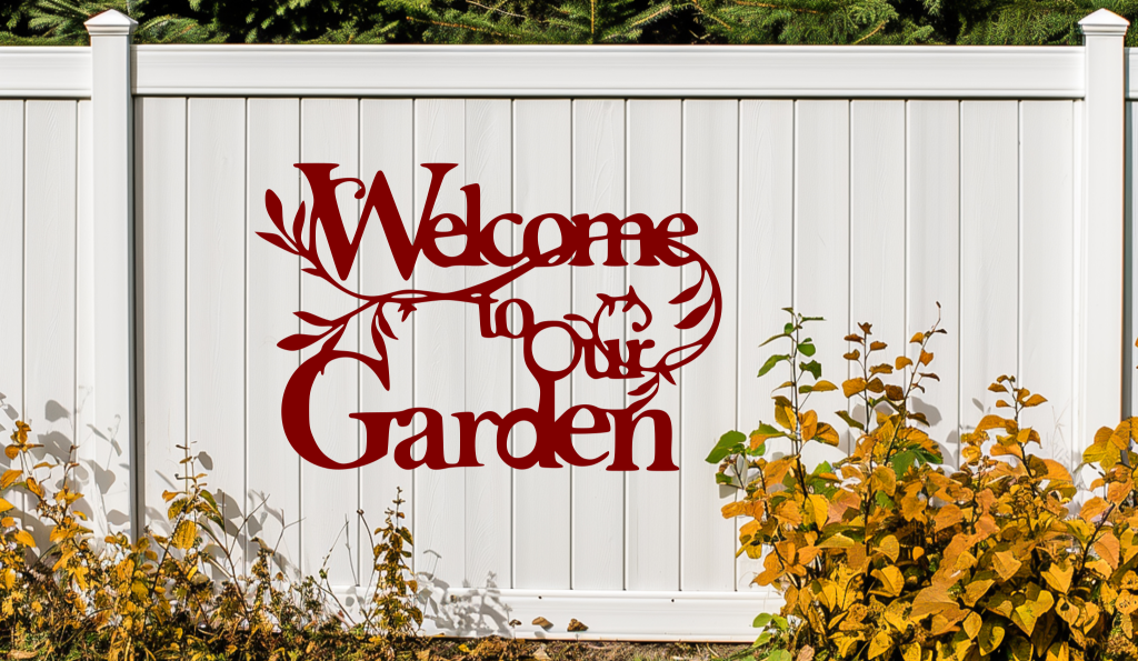 Metal wall art sign featuring the phrase 'Welcome to our Garden' with decorative branches and leaves integrated into the design, finished in Manor Red powder coating. The sign is mounted on a white fence, adding a welcoming touch to an outdoor garden space.