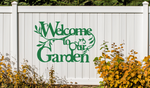 Metal wall art sign featuring the phrase 'Welcome to our Garden' with decorative branches and leaves integrated into the design, finished in Mistletoe powder coating. The sign is mounted on a white fence, adding a welcoming touch to an outdoor garden space