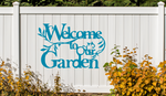 Metal wall art sign featuring the phrase 'Welcome to our Garden' with decorative branches and leaves integrated into the design, finished in Wedgwood Satin powder coating. The sign is mounted on a white fence, adding a welcoming touch to an outdoor garden space.