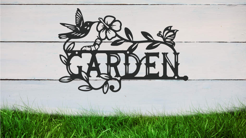 Metal wall art featuring the word 'GARDEN' with a decorative design of leaves, flowers, and a hummingbird, finished in Berry Grey Gloss powder coating, mounted on a white wooden wall above a patch of green grass, adding a welcoming and natural touch to outdoor garden spaces.