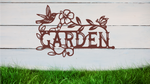 Metal wall art featuring the word 'GARDEN' with a decorative design of leaves, flowers, and a hummingbird, finished in Rust Patina powder coating, mounted on a white wooden wall above a patch of green grass, adding a welcoming and natural touch to outdoor garden spaces