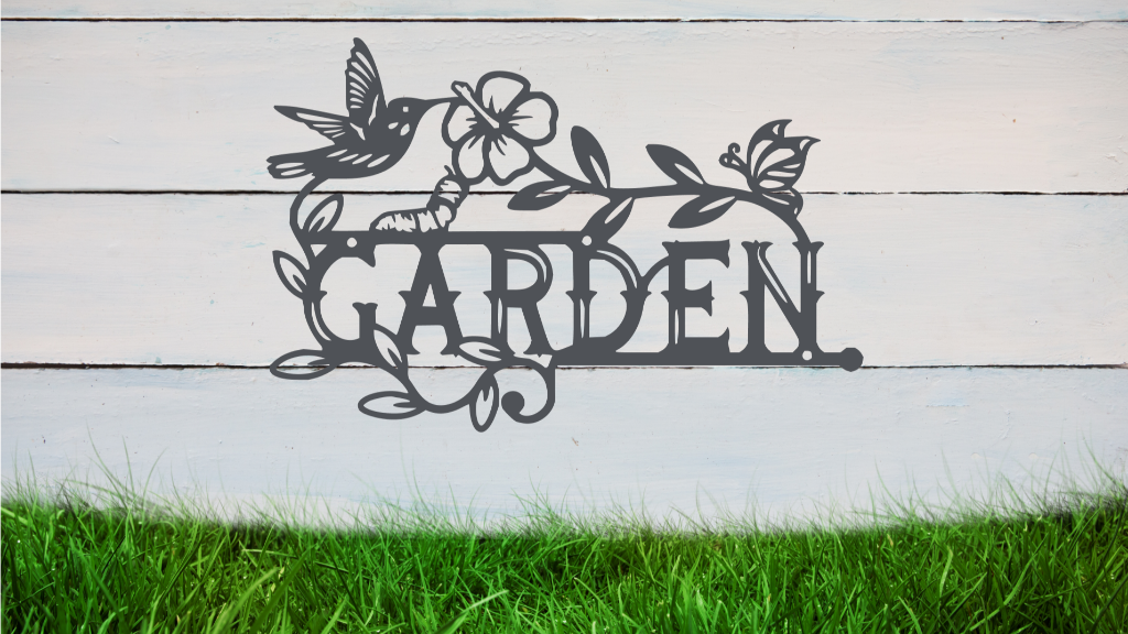 Metal wall art featuring the word 'GARDEN' with a decorative design of leaves, flowers, and a hummingbird, finished in Berry Grey Gloss powder coating, mounted on a white wooden wall above a patch of green grass, adding a welcoming and natural touch to outdoor garden spaces.