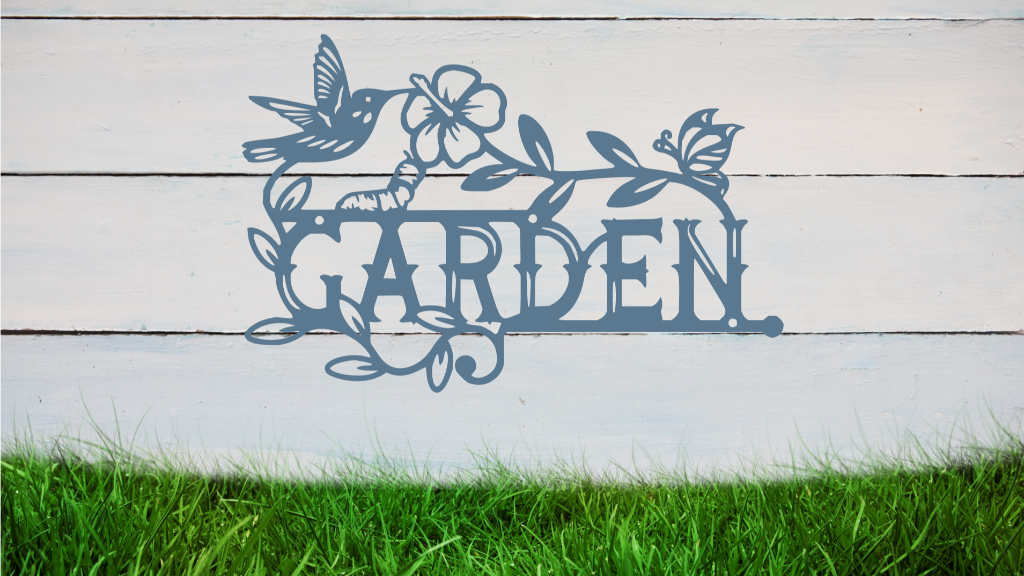 Metal wall art featuring the word 'GARDEN' with a decorative design of leaves, flowers, and a hummingbird, finished in Wedgwood Satin powder coating, mounted on a white wooden wall above a patch of green grass, adding a welcoming and natural touch to outdoor garden spaces.