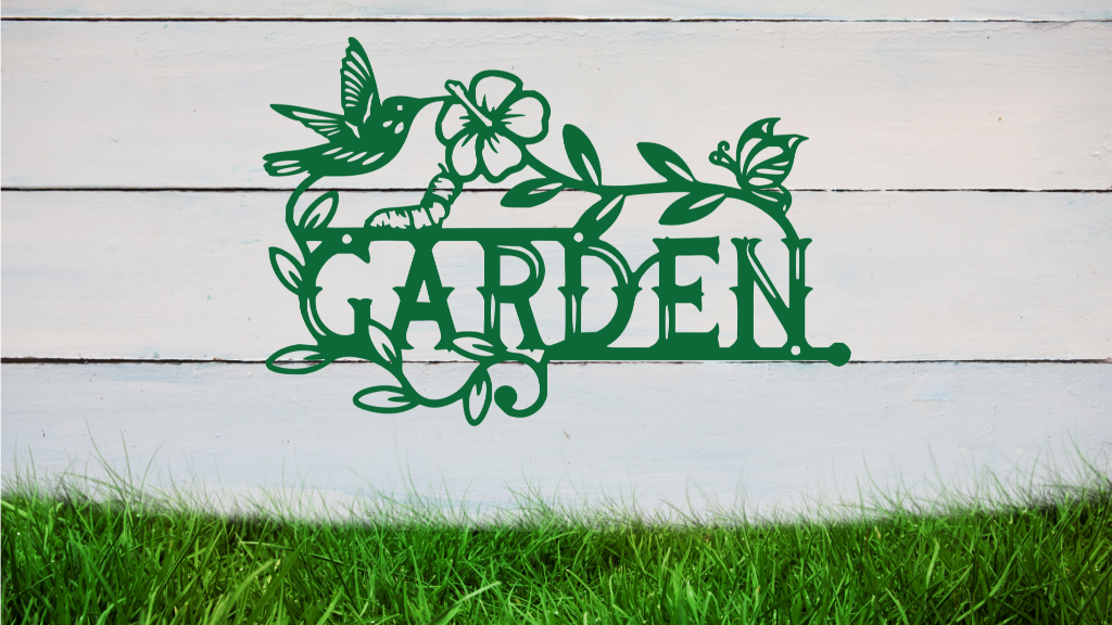 Metal wall art featuring the word 'GARDEN' with a decorative design of leaves, flowers, and a hummingbird, finished in Mistletoe powder coating, mounted on a white wooden wall above a patch of green grass, adding a welcoming and natural touch to outdoor garden spaces.