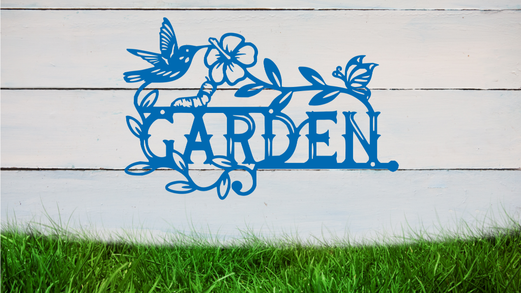 Metal wall art featuring the word 'GARDEN' with a decorative design of leaves, flowers, and a hummingbird, finished in Wedgwood Satin powder coating, mounted on a white wooden wall above a patch of green grass, adding a welcoming and natural touch to outdoor garden spaces.