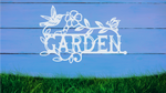 Metal wall art featuring the word 'GARDEN' with a decorative design of leaves, flowers, and a hummingbird, finished in Bright White Gloss powder coating, mounted on a white wooden wall above a patch of green grass, adding a welcoming and natural touch to outdoor garden spaces.