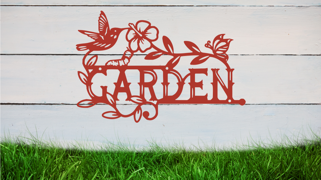 Metal wall art featuring the word 'GARDEN' with a decorative design of leaves, flowers, and a hummingbird, finished in Manor Red powder coating, mounted on a white wooden wall above a patch of green grass, adding a welcoming and natural touch to outdoor garden spaces.