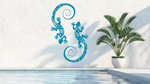 Metal wall art featuring two geckos with intricate detailing on their bodies, finished in Wedgwood Satin powder coating, mounted on a light wall near a poolside, adding a tropical and playful touch to the outdoor space.