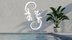 Metal wall art featuring two geckos with intricate detailing on their bodies, finished in Bright White Gloss powder coating, mounted on a light wall near a poolside, adding a tropical and playful touch to the outdoor space.