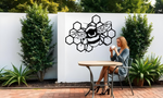 Metal wall art featuring a detailed bee resting on a honeycomb design, finished in Night Sky Matt Black powder coating, mounted on a white outdoor wall. The art adds a charming and nature-inspired focal point to a garden or patio area, where a woman enjoys a cup of coffee while seated at a table.