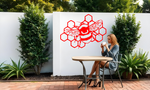 Metal wall art featuring a detailed bee resting on a honeycomb design, finished in Signal Red powder coating, mounted on a white outdoor wall. The art adds a charming and nature-inspired focal point to a garden or patio area, where a woman enjoys a cup of coffee while seated at a table.