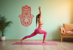 Decorative Hamsa hand metal wall art in Signal Red, mounted on a pastel green wall next to a yoga enthusiast in a warrior pose.