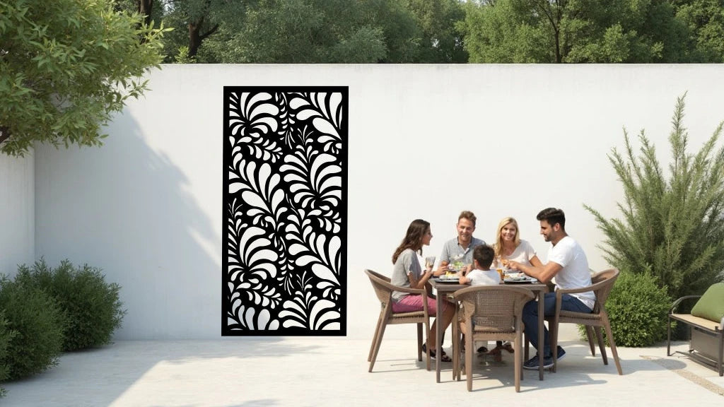 The 'Jungle Mirage' metal privacy screen featuring an intricate swirling leaf pattern, finished in Night Sky Matt Black powder coating. The screen is mounted on a white wall in an outdoor patio area, where a group of five people are gathered around a dining table enjoying a meal together, adding elegance and subtle privacy to the space