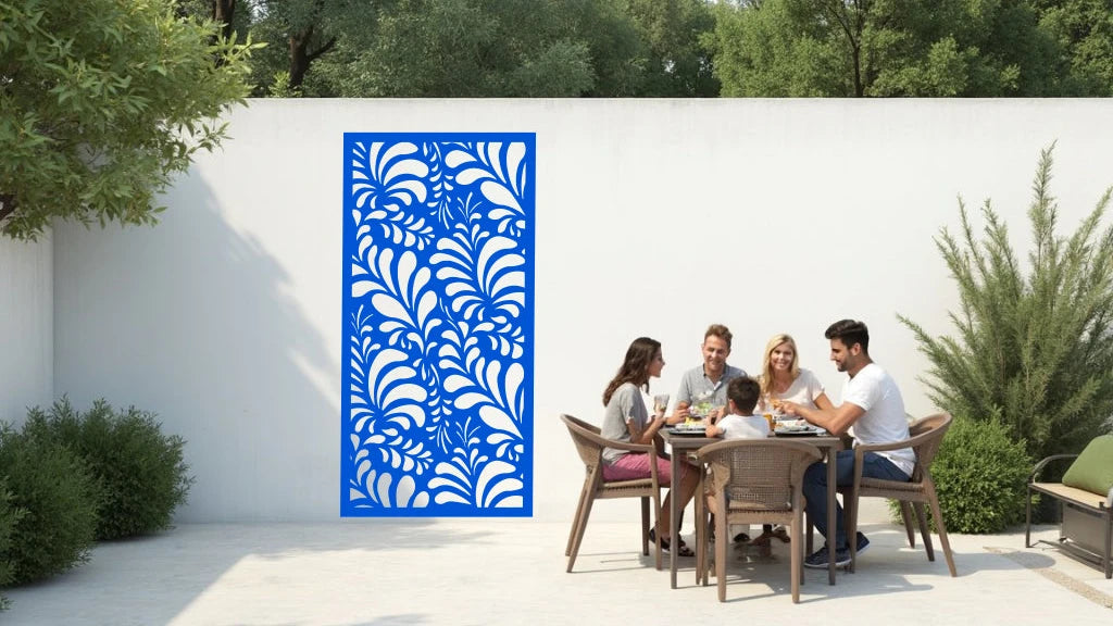 The 'Jungle Mirage' metal privacy screen featuring an intricate swirling leaf pattern, finished in Blaze Blue Gloss powder coating. The screen is mounted on a white wall in an outdoor patio area, where a group of five people are gathered around a dining table enjoying a meal together, adding elegance and subtle privacy to the space