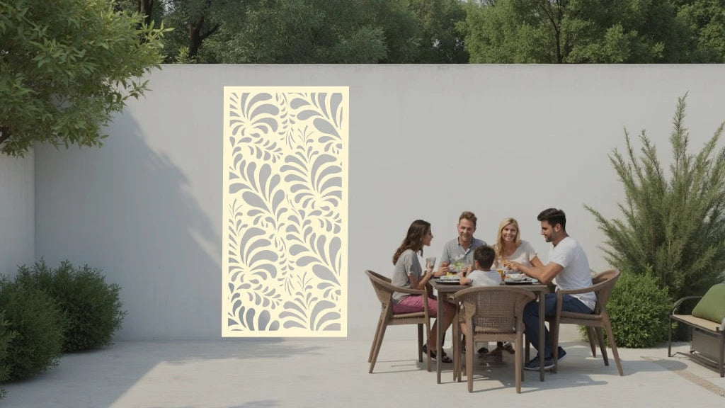 The 'Jungle Mirage' metal privacy screen featuring an intricate swirling leaf pattern, finished in Canola Cream powder coating. The screen is mounted on a white wall in an outdoor patio area, where a group of five people are gathered around a dining table enjoying a meal together, adding elegance and subtle privacy to the space.