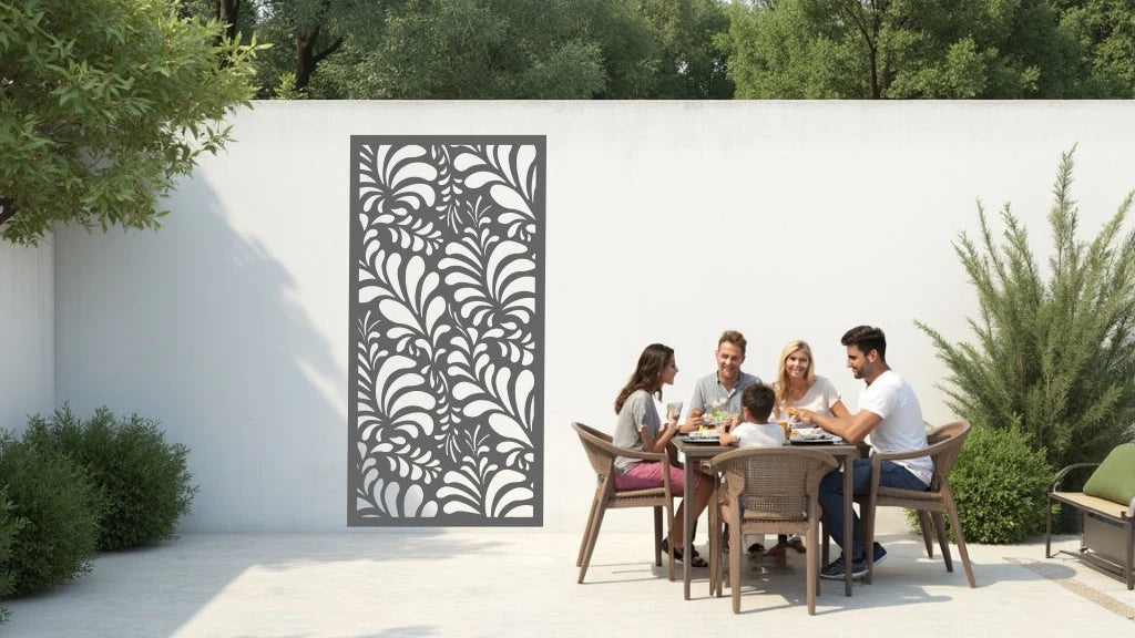 The 'Jungle Mirage' metal privacy screen featuring an intricate swirling leaf pattern, finished in Berry Grey Gloss powder coating. The screen is mounted on a white wall in an outdoor patio area, where a group of five people are gathered around a dining table enjoying a meal together, adding elegance and subtle privacy to the space