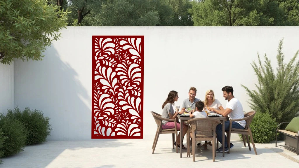 The 'Jungle Mirage' metal privacy screen featuring an intricate swirling leaf pattern, finished in Manor Red powder coating. The screen is mounted on a white wall in an outdoor patio area, where a group of five people are gathered around a dining table enjoying a meal together, adding elegance and subtle privacy to the space