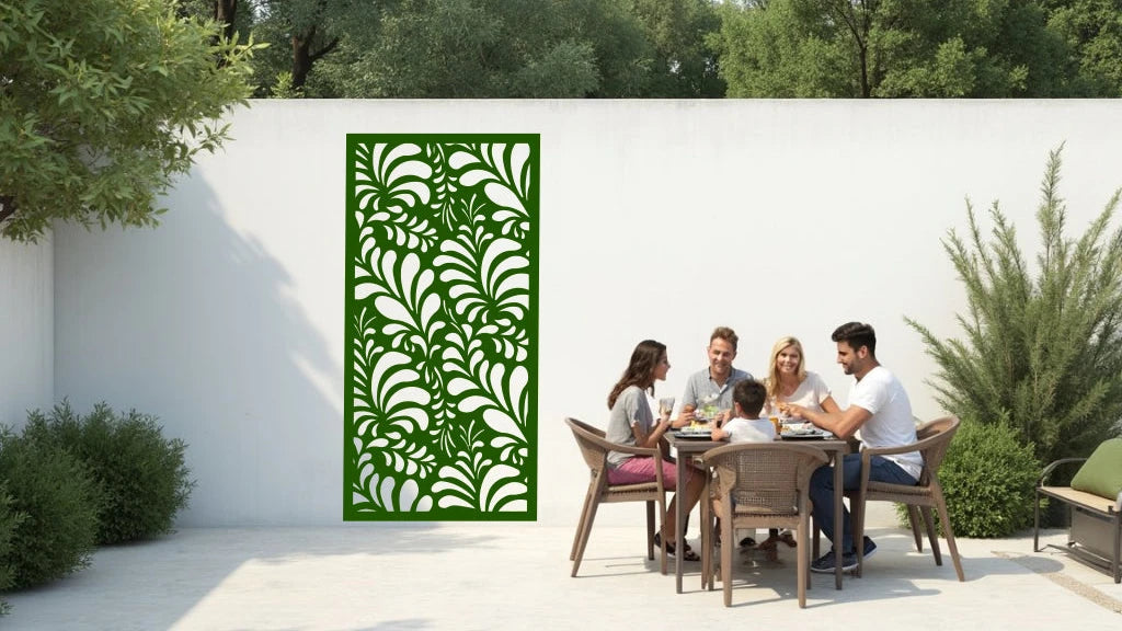 The 'Jungle Mirage' metal privacy screen featuring an intricate swirling leaf pattern, finished in Mistletoe powder coating. The screen is mounted on a white wall in an outdoor patio area, where a group of five people are gathered around a dining table enjoying a meal together, adding elegance and subtle privacy to the space