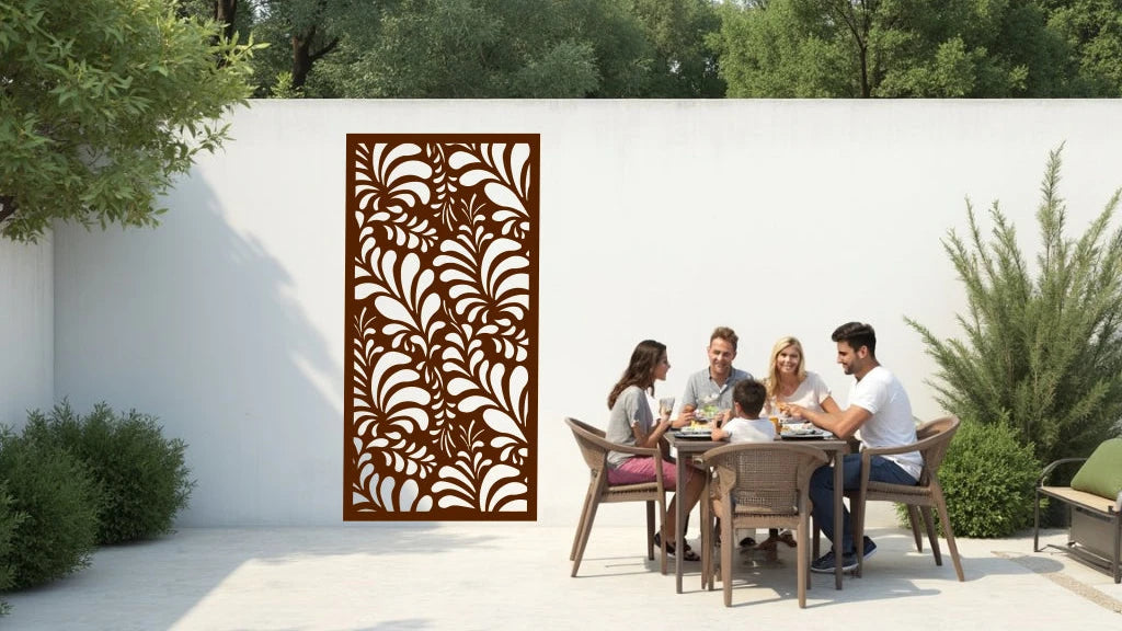 The 'Jungle Mirage' metal privacy screen featuring an intricate swirling leaf pattern, finished in Rust Patina powder coating. The screen is mounted on a white wall in an outdoor patio area, where a group of five people are gathered around a dining table enjoying a meal together, adding elegance and subtle privacy to the space