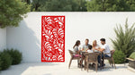 The 'Jungle Mirage' metal privacy screen featuring an intricate swirling leaf pattern, finished in Signal Red powder coating. The screen is mounted on a white wall in an outdoor patio area, where a group of five people are gathered around a dining table enjoying a meal together, adding elegance and subtle privacy to the space